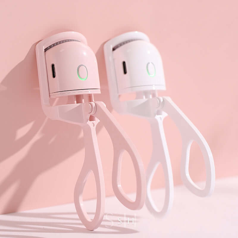 Portable electric heated eyelash curlers in pink and white colors against a soft pink background.