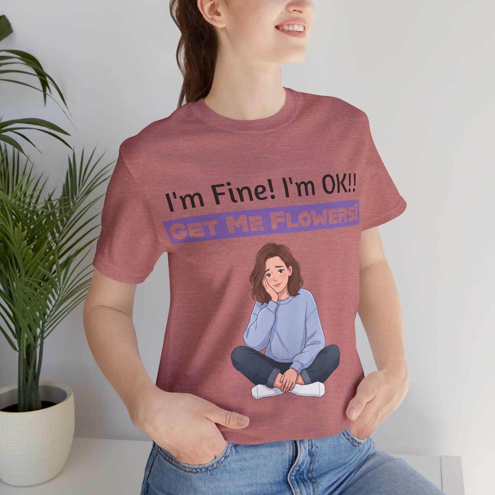 A woman wearing a dusty rose t-shirt featuring a cartoon girl and the text I'm Fine! I'm OK!! GET ME FLOWERS!