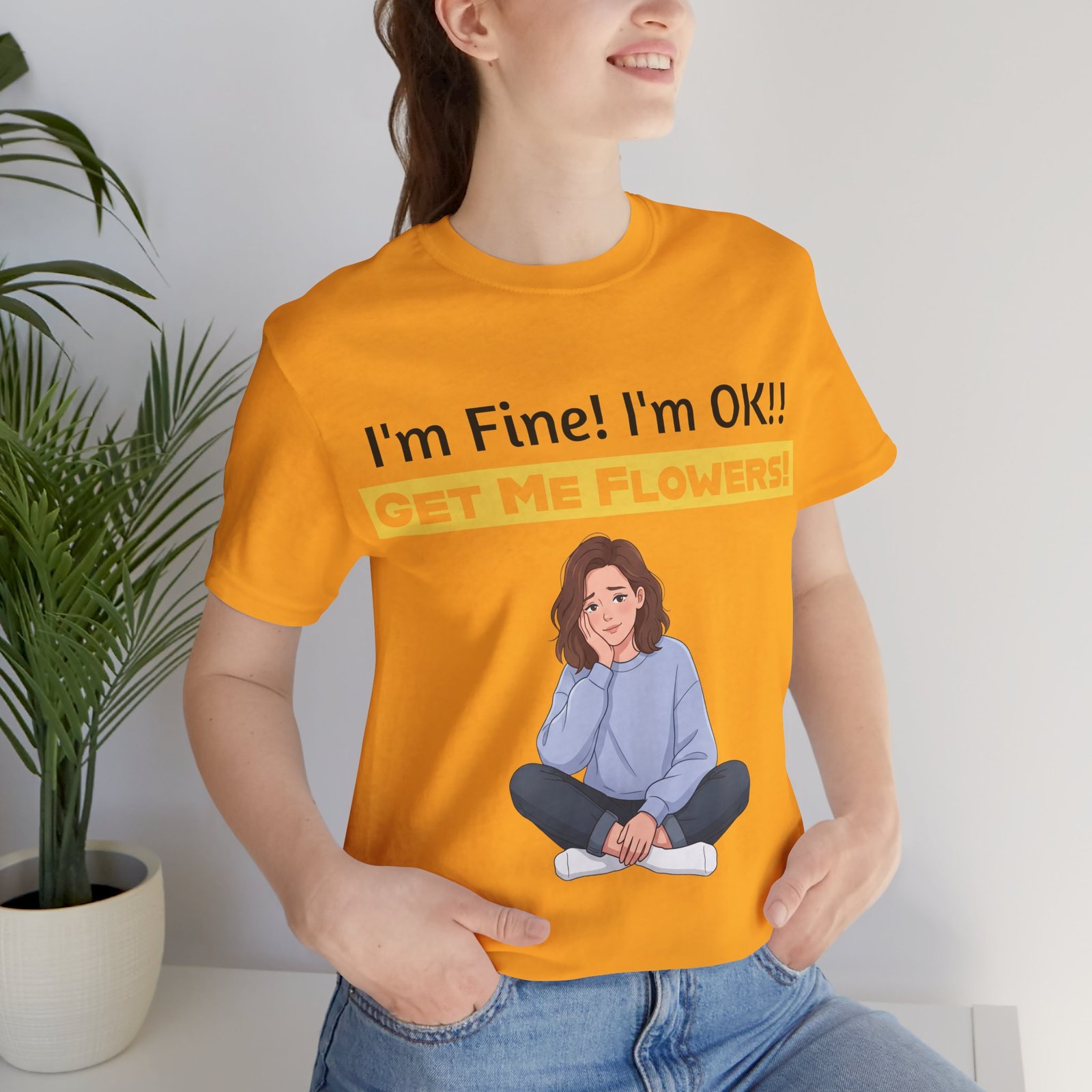 A woman wearing an orange T-shirt with an illustration of a girl and text: I'm Fine! I'm OK!! GET ME FLOWERS!