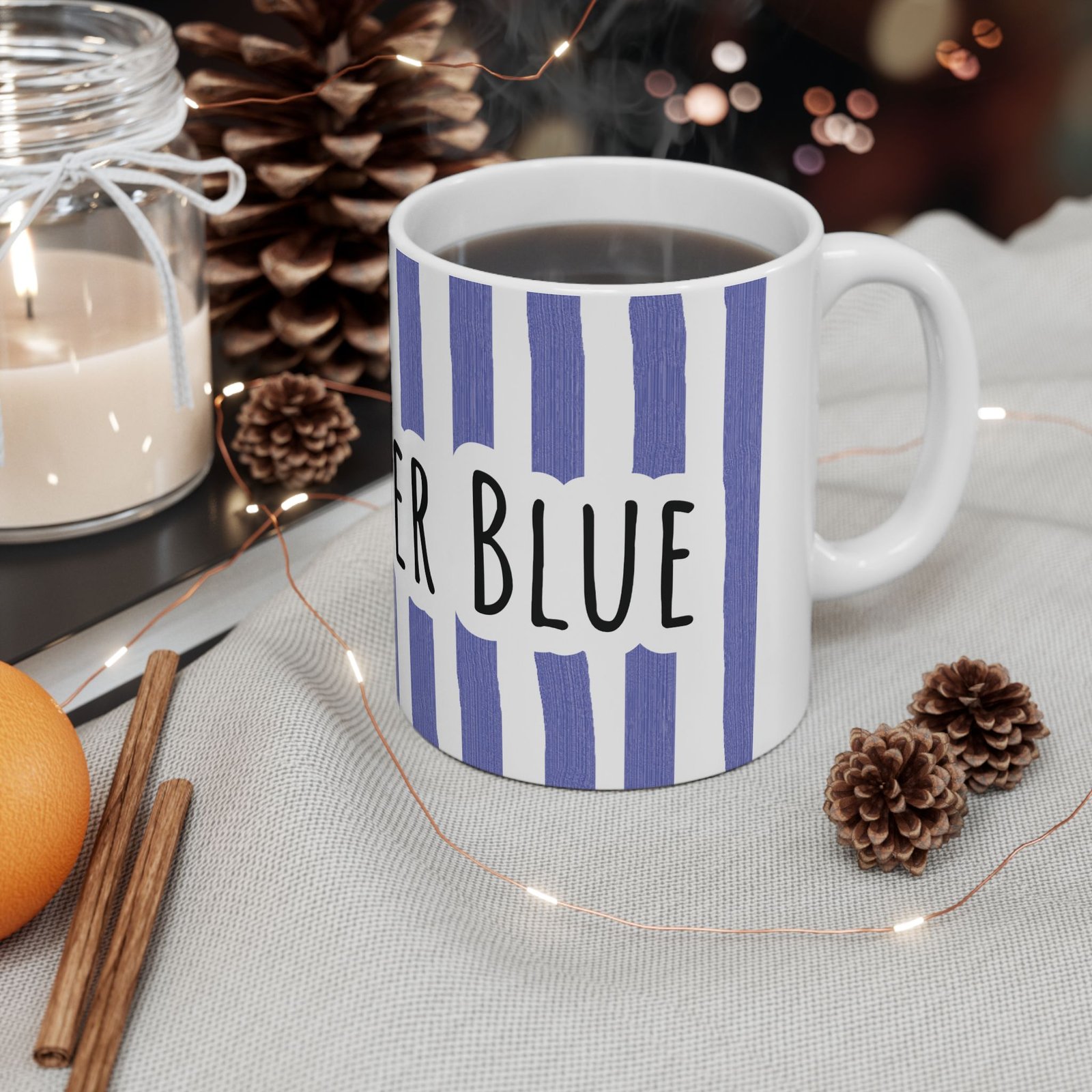 Periwinkle striped ceramic mug with hand-lettered 'Stay Under Blue' text, set among pinecones and a candle.