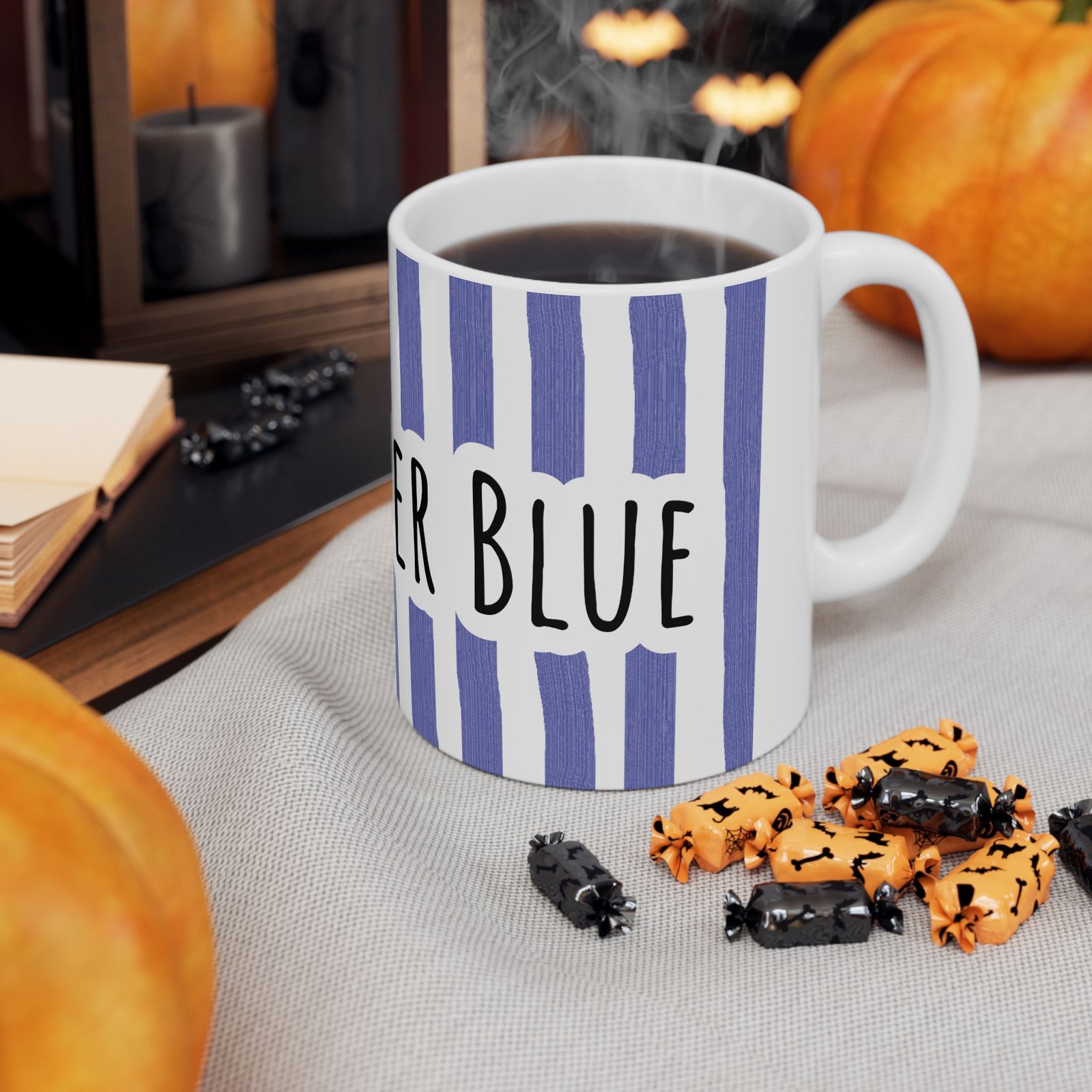 Periwinkle striped ceramic mug with hand-lettered 'Stay Under Blue' text sitting on a cloth next to orange candy.