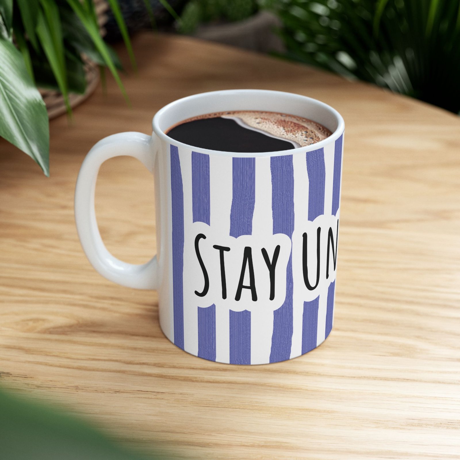 Periwinkle striped ceramic mug with hand-lettered 'STAY UNDER BLUE' text sitting on a wooden surface next to greenery.