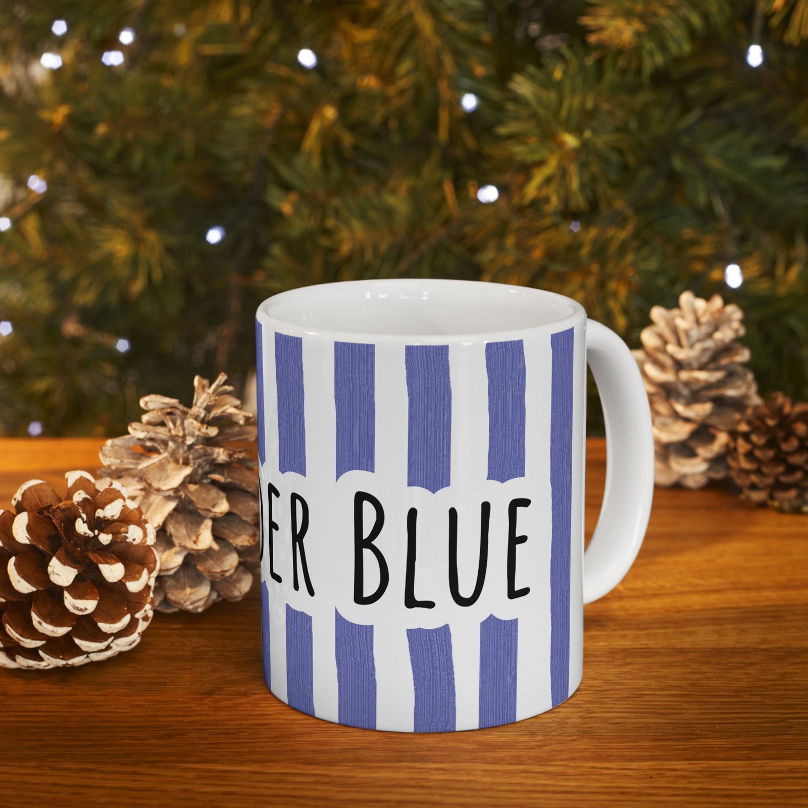 Periwinkle striped ceramic mug with hand-lettered 'Stay Under Blue' text sitting on a wooden surface with pinecones.