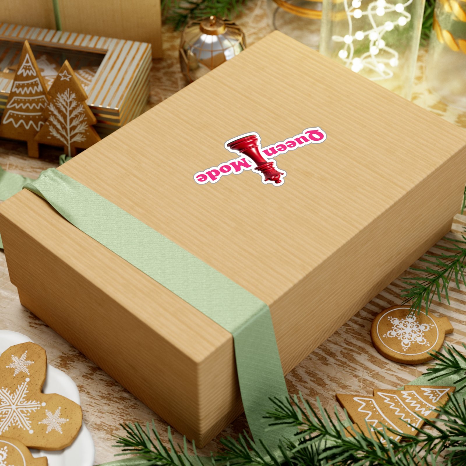 Queen Mode red chess queen sticker with pink text on a light wood gift box decorated with greenery and cookies.