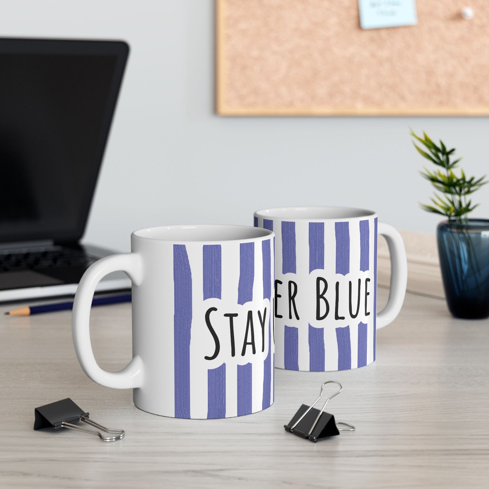 Two periwinkle striped ceramic mugs with hand-lettered 'Stay Under Blue' text on a wooden desk near a laptop.
