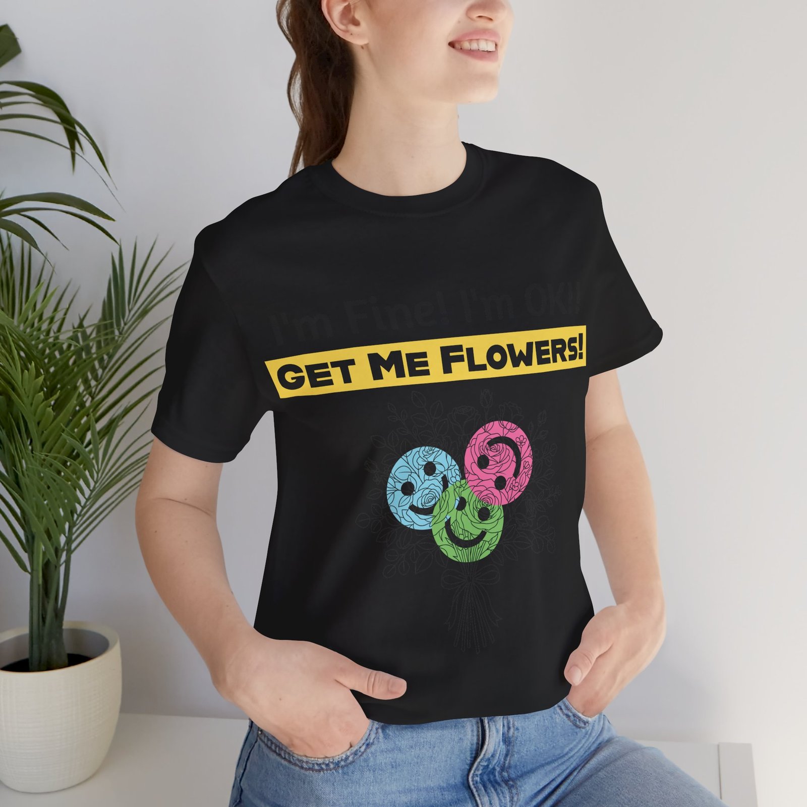 Woman wearing a black crew-neck tee with 'I'm Fine! I'm OK!! GET ME FLOWERS!' text and colorful smiley face blooms.