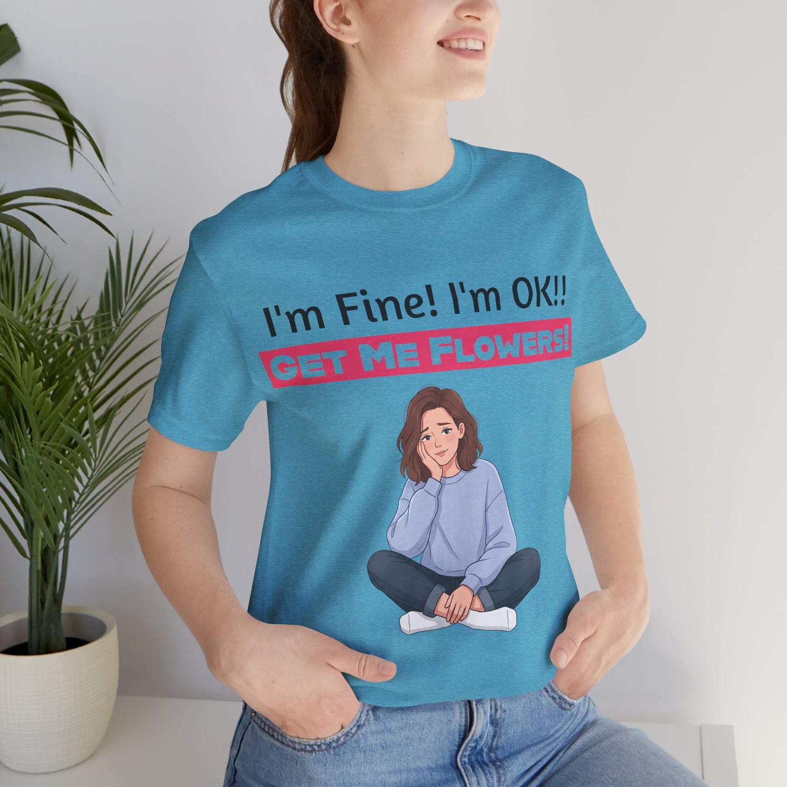 Woman wearing a blue graphic t-shirt that says "I'm Fine! I'm OK!! GET ME FLOWERS!" with an illustration of a seated girl.