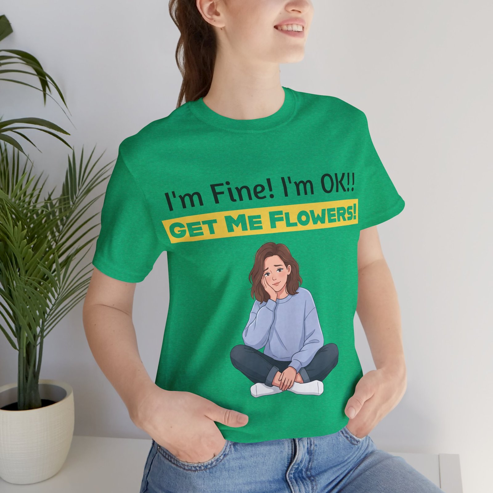 Woman wearing a green crew neck t-shirt with a funny graphic of a girl sitting and the text I'm Fine! I'm OK!! GET ME FLOWERS!