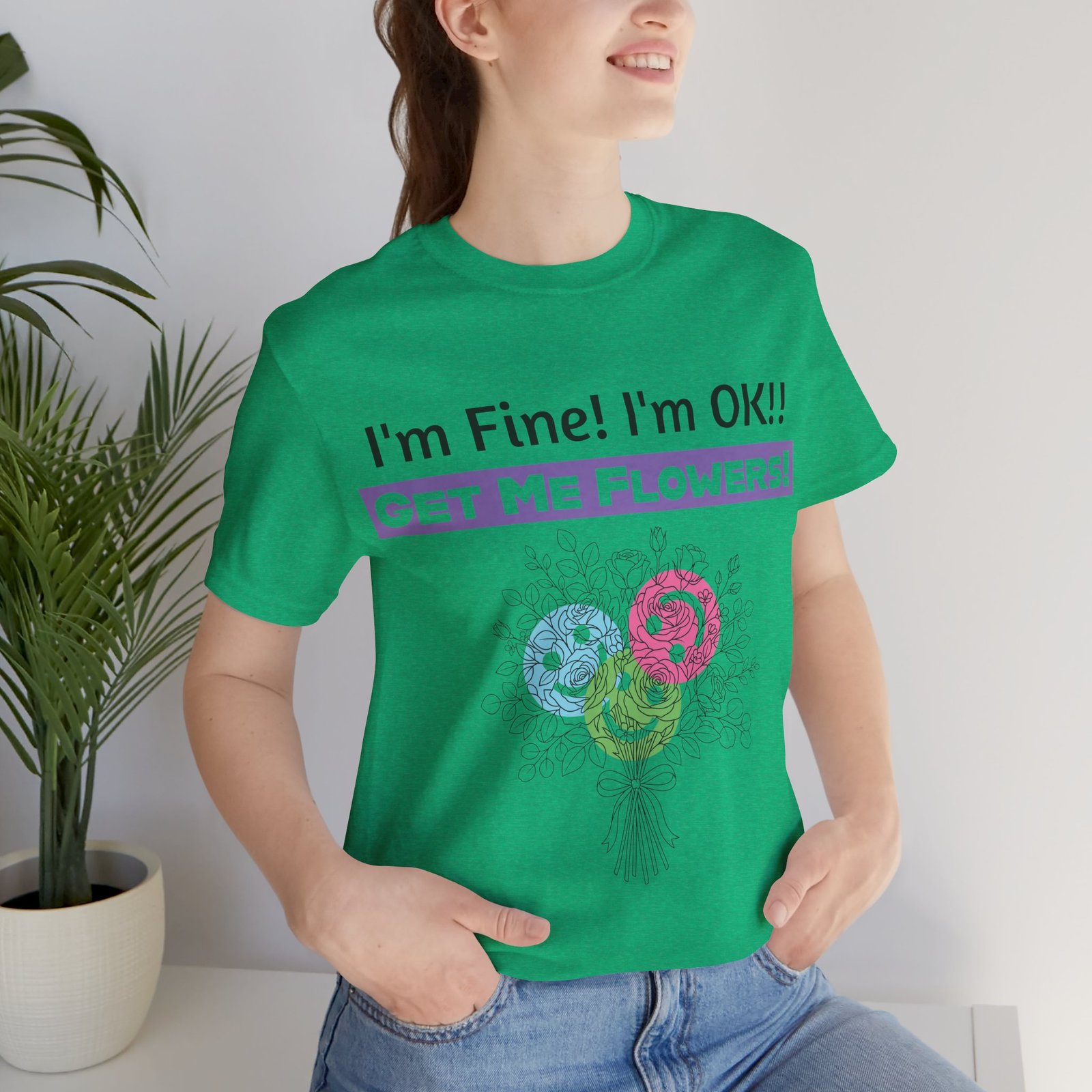 Woman wearing a green graphic t-shirt featuring the sarcastic text "I'm Fine! I'm OK!! GET ME FLOWERS!" and colorful blooms.
