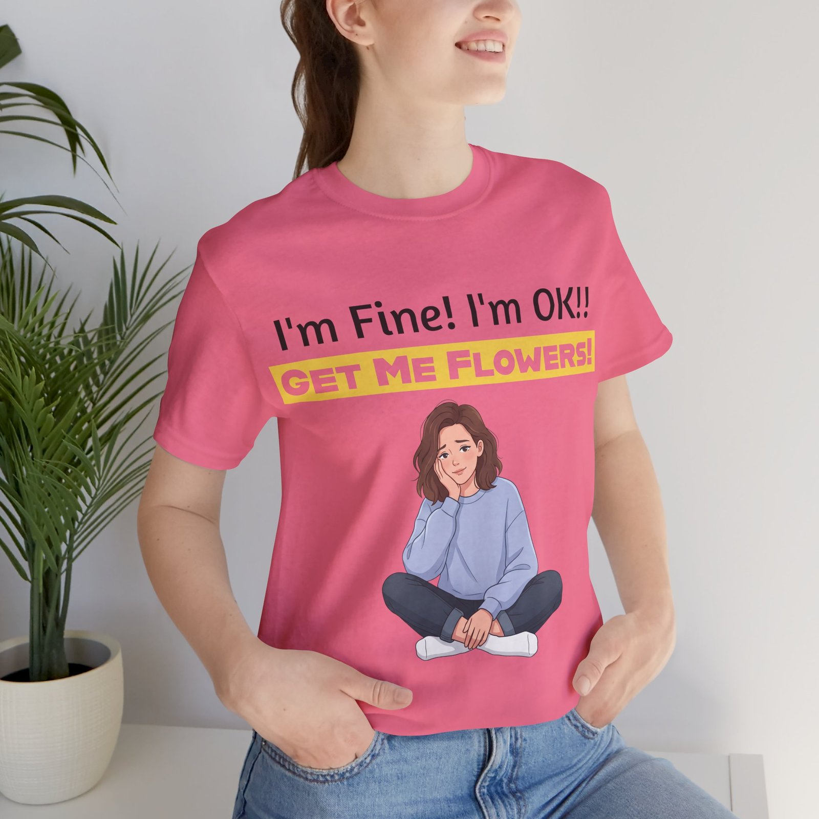 Woman wearing a pink 'I'm Fine! I'm OK!! GET ME FLOWERS!' funny T-shirt with a cartoon girl illustration.