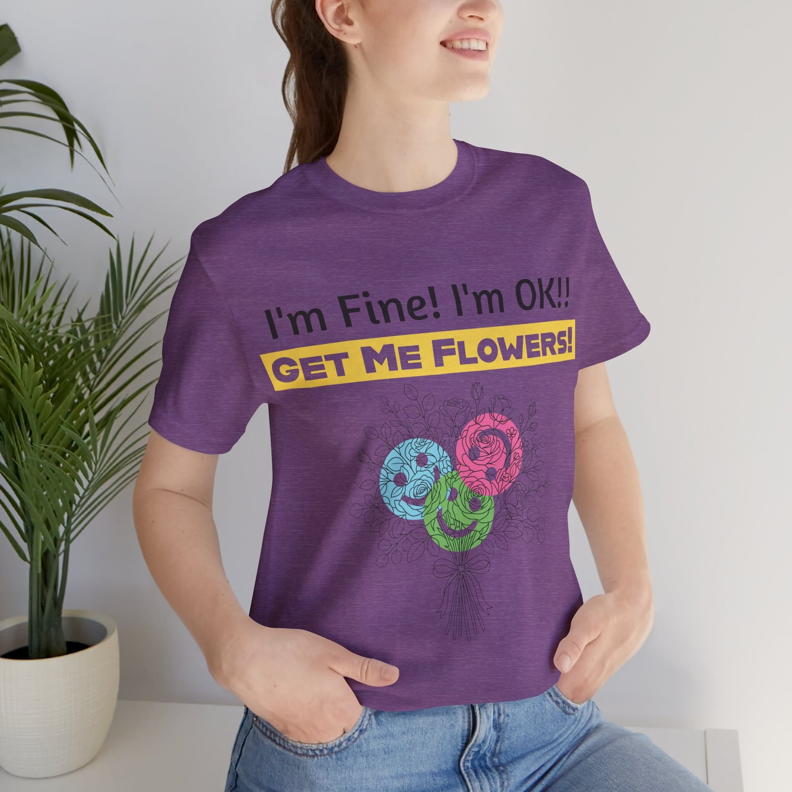 Woman wearing a purple Bella+Canvas t-shirt with 'I'm Fine! I'm OK!! GET ME FLOWERS!' text and colorful flower graphic.