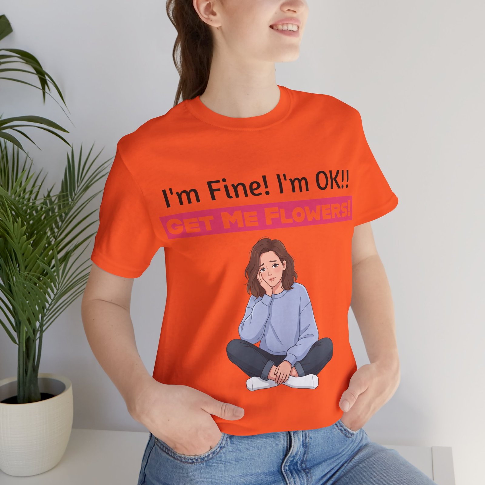 Woman wearing an orange funny t-shirt with 'I'm Fine! I'm OK!! GET ME FLOWERS!' text and a cartoon graphic.