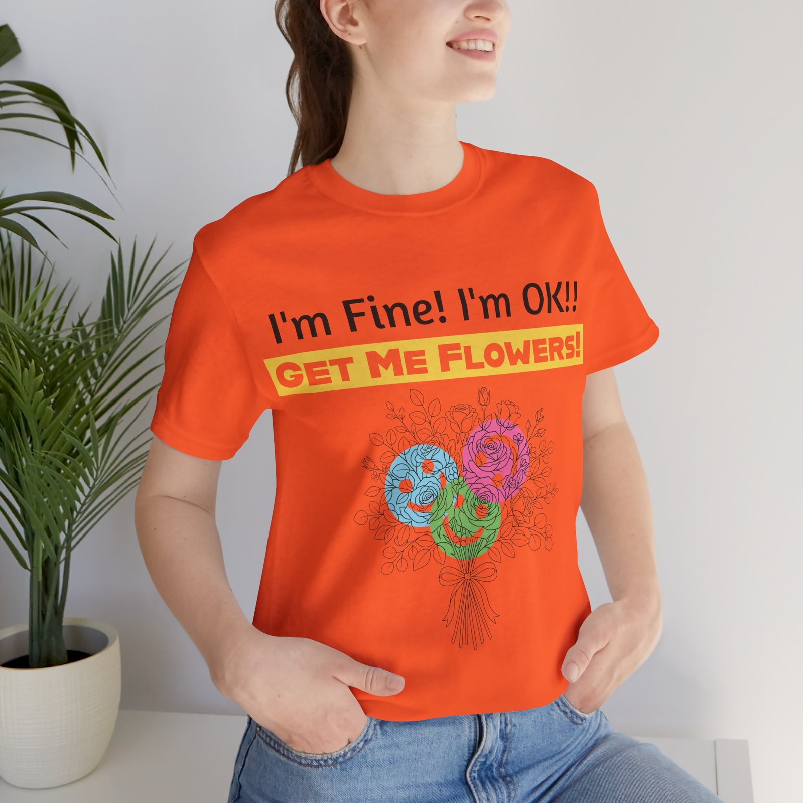 Woman wearing an orange t-shirt with 'I'm Fine! I'm OK!! GET ME FLOWERS!' text and colorful abstract flower graphic.